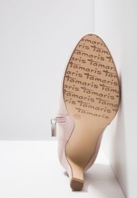Botte à talon en daim rose reposant semelle vers le haut contre un coin blanc, semelle embossée avec le logo "Tamaris" répété et la taille 37 marquée.