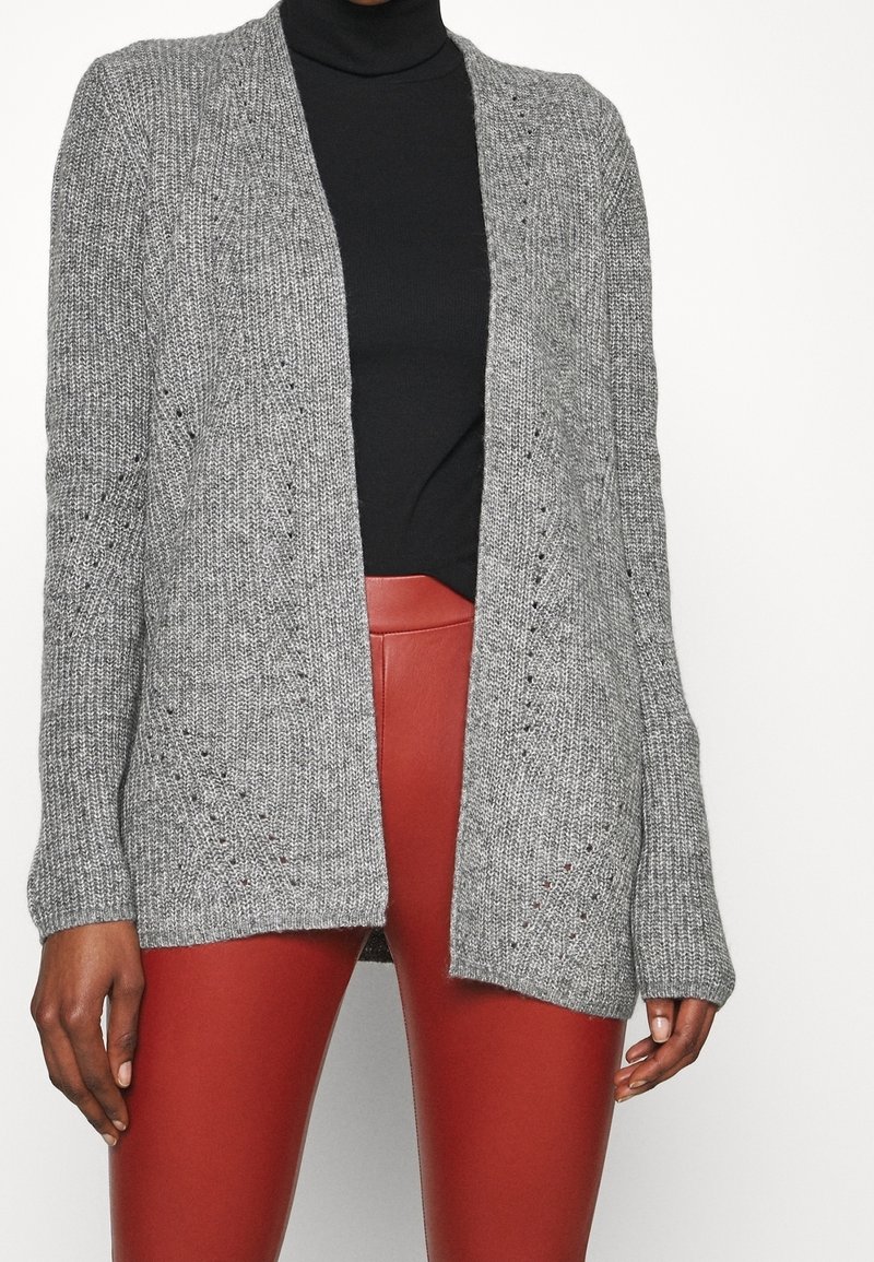 Cardigan gris en maille avec un devant ouvert, des poignets côtelés et des trous décoratifs. Associé à un pantalon rouge en fausse cuir ajusté.