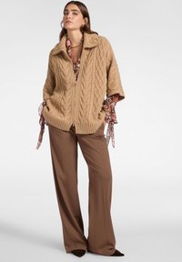 Maglione beige a maglia cable con chiusura a zip e colletto ampio, abbinato a pantaloni marroni larghi. Presenta una camicetta con motivo floreale sotto.
