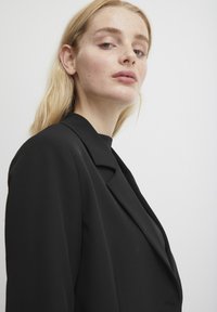 Zwarte blazer met een gestructureerd ontwerp, met opgesneden revers en subtiele schoudervulling. De stof lijkt glad en heeft een getailleerde pasvorm.