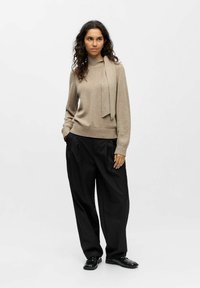 Pull en tricot beige avec un détail à nouer au niveau du col, assorti à un pantalon noir ample et des chaussures noires brillantes.