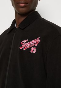 Giacca in pile nera con chiusura a zip, caratterizzata da un logo "Tommy" ricamato in rosso e bianco e un design con colletto classico.