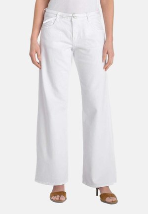 Donna che indossa pantaloni bianchi a gamba larga con tasche frontali e sandali con tacco marroni aperti, in piedi davanti a uno sfondo semplice.