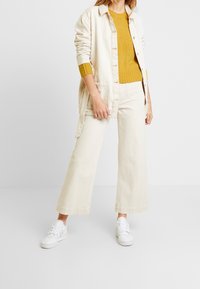 Béžová oversized košile, hořčicový svetr a široké béžové kalhoty, doplněné bílými teniskami. Bavlněný materiál s volným střihem.