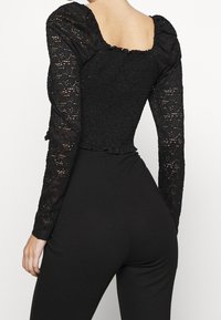 Svart bodysuit med långa spetsärmar, med en figurnära smockad byst, fyrkantig halsringning och volangdetalj i fållen.