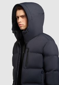 Sötét tengeri kék puffermellény nagy kapucnival, steppelt dizájnnal, elülső cipzárral és egy oldalsó cipzáras zsebbel a funkciók bővítése érdekében.