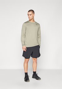 Helles olivfarbenes Langarm-Sportshirt mit schwarzem Logo, kombiniert mit schwarzen Shorts und schwarzen Sportschuhen. Das Modell steht vor einem neutralen Hintergrund.
