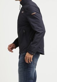 Mörkblå zip-up jacka med hög krage, sidfickor och knappmanchetter. Har en logotyp på ärmen och en smidig, stilren textur.