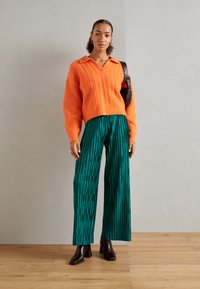 Sweater de punto trenzado naranja con cuello, combinado con pantalones de pierna ancha verdes con pliegues y rayas verticales. Botines de tacón marrones.