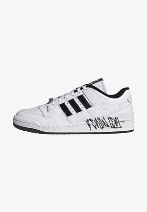 Baskets basses blanches Adidas avec des bandes noires, une doublure intérieure noire et un texte noir style graffiti sur la semelle intermédiaire.
