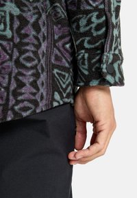 Chaqueta de forro polar con patrones abstractos en negro, púrpura y verde azulado; cuenta con un puño abotonado y tejido texturizado; combinada con pantalones oscuros.
