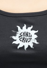 Μαύρο ριμπ ύφασμα με λευκό σχέδιο σε σχήμα ήλιου και το κείμενο "Santa Cruz" σε στυλιζαρισμένα κεφαλαία γράμματα στο κέντρο του στήθους.