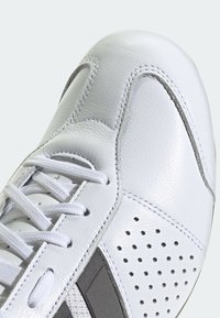 Detail bílých tenisek, zobrazující špičku s perforacemi, černobílou tkanou látkou a bílými tkaničkami na hladkých kožených dílech.