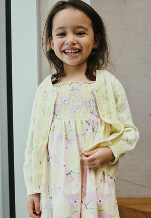 Jeune fille souriante aux cheveux foncés portant une robe fleurie jaune pastel et un cardigan tricoté jaune clair, debout à l’intérieur près d’une fenêtre.