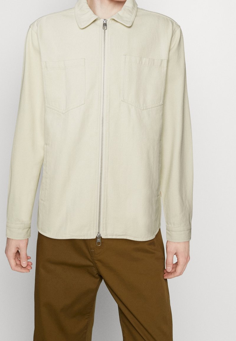 Mann trägt eine beigefarbene Jacke mit Reißverschluss und zwei Brusttaschen sowie braune Hose, stehend vor einem schlichten weißen Hintergrund.