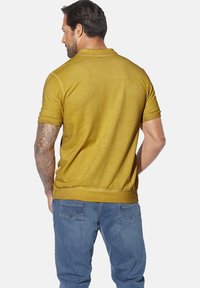 Short-sleeve polo shirt in mustard yellow, made of soft material. Features a ribbed collar and hem, with a smooth texture and fitted design.