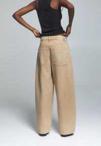 Beżowe szerokie spodnie dżinsowe, z wysokim stanem, dwoma tylnymi kieszeniami i surowym wykończeniem. Stylizowane z czarnym topem na ramiączkach.