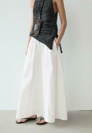 Personne portant un haut tricoté noir sans manches avec un pantalon blanc à jambes larges, des tongs noires et un petit collier avec une pochette marron.