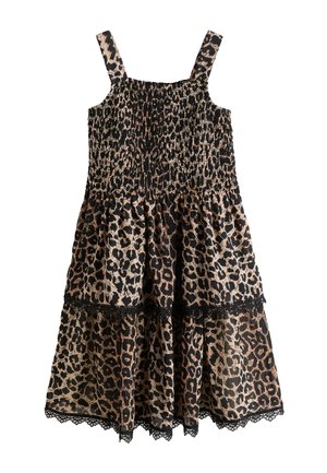 Ragazza con lunghi capelli castani che indossa un vestito senza maniche con stampa leopardata e sandali neri, in piedi su uno sfondo chiaro e uniforme.