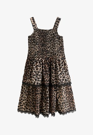 Meisje met lang bruin haar, gekleed in een mouwloze jurk met luipaardprint en zwarte sandalen, staand tegen een effen lichte achtergrond.