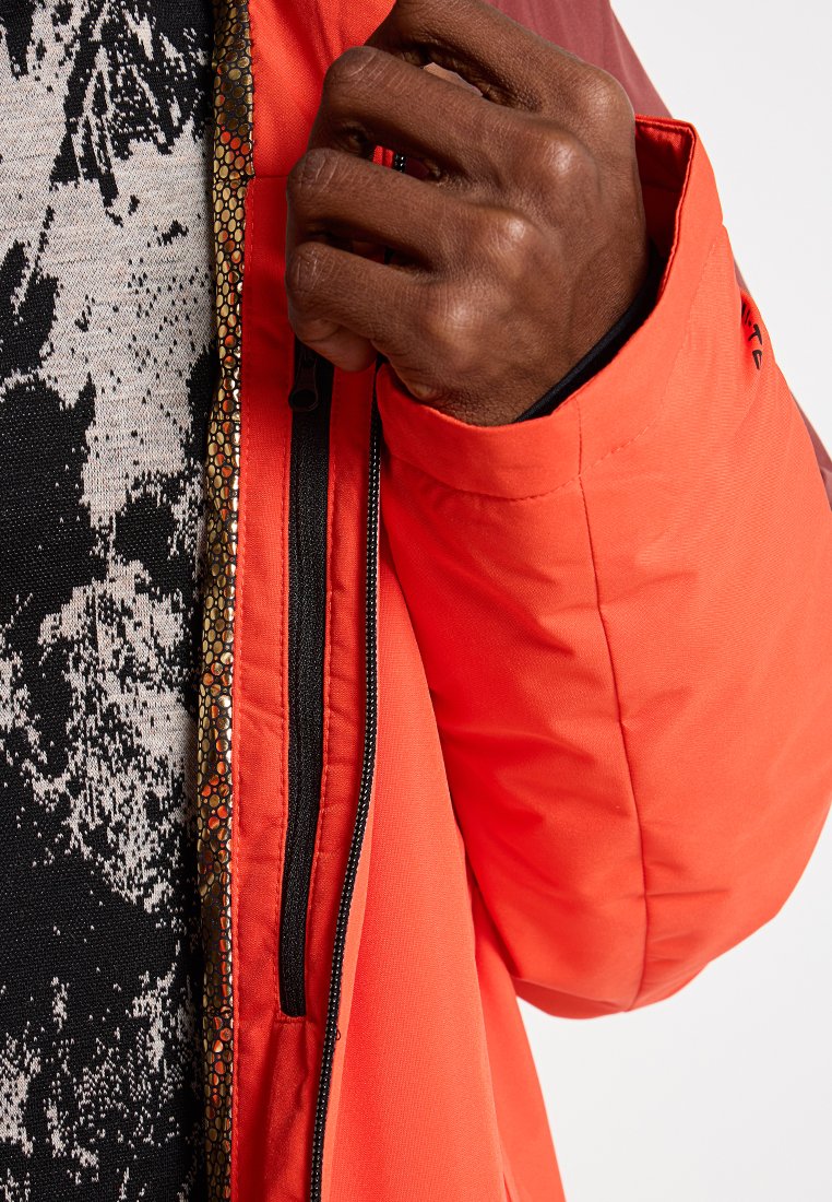 Hand mit dunkler Hautfarbe, die den Reißverschluss einer orangefarbenen Jacke anpasst, die über einem schwarz-weißen Mustersweater getragen wird.