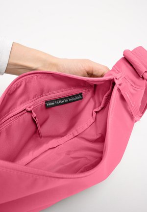 Hand hält eine offene pinke Tasche, die eine innere Reißverschlusstasche mit einem Etikett zeigt, auf dem "VON MÜLL ZU SCHATZ" steht.