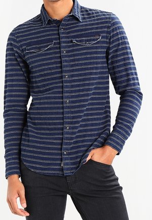 Camicia button-up blu navy con righe orizzontali bianche, due tasche sul petto e colletto. Il tessuto sembra strutturato con una leggera elasticità.