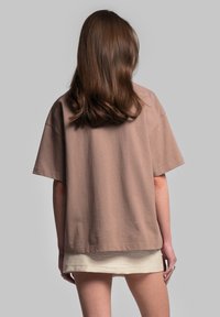 Overdimensioneret t-shirt med korte ærmer i muted brun bomuld, med rund hals og lige kant. Båret over et lyst beige plagg.