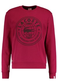 Sweat-shirt rouge à manches longues avec logo Lacoste noir et texte « All Around The World » sur le devant, poignets et ourlet côtelés.