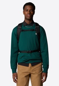 Green sweatshirt with a textured finish, layered over a black collared shirt. Includes a black harness-style strap. Tan pants below.