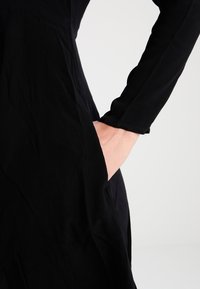 Svart klänning med långa ärmar och sidfickor, tillverkad av mjukt tyg med en slät textur, närbild på fickdetaljen.