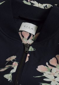 Veste bombardier bleu marine avec motif floral rose ; comprend une fermeture éclair noire et un col côtelé ; l'intérieur est doublé et doux. Étiquette de la marque visible.