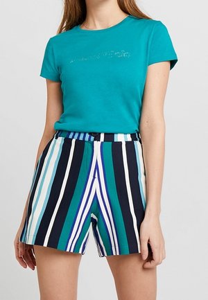 Kvinde iført en teal t-shirt med korte ærmer og højtaljede shorts med lodrette striber i blå, hvid og grøn på en ensfarvet baggrund.