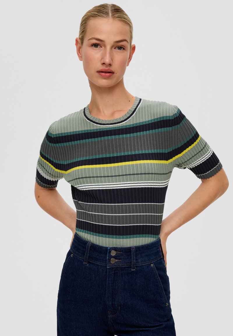 Jonge vrouw met blond haar in een gestreepte ribbeltrui met korte mouwen en hoograndige donkere spijkerbroek, met haar handen op haar heupen.