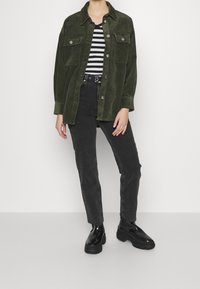 Zelená kordová bunda přes černobíle pruhovanou košili, v kombinaci s černými džíny a silnými černými botami, stojící na bílém pozadí.