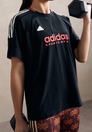 Persona vistiendo ropa deportiva negra de Adidas levanta una pesa negra, mostrando torso superior y mallas estampadas con diseño de palmeras.