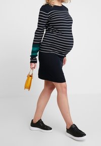 Těhotenské šaty s námořnickými a bílými pruhy, ke kolenům, nošeny s černými teniskami, doplněny malou žlutou kabelkou. Texturované pletené rukávy.