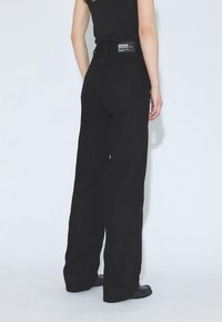 Jeans negros de pierna ancha hechos de mezclilla, con cintura alta y detalle de bolsillo trasero. La cinturilla muestra una etiqueta con el logo e información de cuidado.