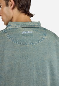 Chaqueta de denim azul claro con un collar clásico y una costura circular en la parte trasera. Logo "Pegador" bordado cerca del cuello en blanco.