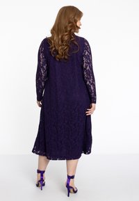 Paarse kanten jurk met lange kanten mouwen en een uitlopende rok. Bevat een getextureerd bloemenpatroon, gedragen met paarse hoge hakken.