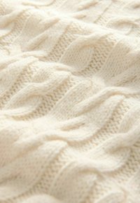 Tissu tricoté en crème texturé présentant des motifs de câble en relief et ondulés. Apparence douce et confortable avec une texture dense et chaude.
