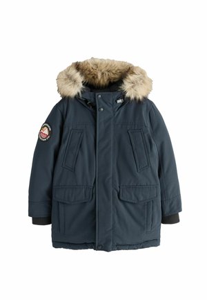 Pojke som bär en mörkblå vinterjacka med pälskantad huva och en tygmärke på ärmen, tittar något uppåt och åt sidan.