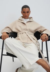 Donna con capelli raccolti seduta su una sedia nera, indossa un cappotto beige con cintura, pantaloni bianchi e scarpe beige, guarda avanti con fiducia.