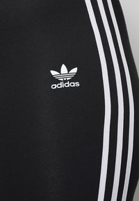 Zwarte sportleggings met drie witte strepen aan de zijkant, met een wit adidas-logo op de onderkant van de voorkant. Gladde, rekbare stof.