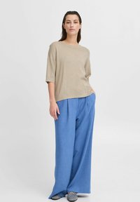 Beige volný pletený top s krátkými rukávy, kombinovaný s modrými širokými kalhotami. Měkčená textura a lehký materiál. Šedé boty dotvářejí vzhled.