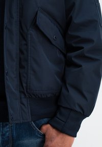 Marineblaue Bomberjacke aus strukturiertem Material, mit Reißverschluss, gerippten Bündchen und Saum sowie einer seitlichen Tasche mit Druckknopfverschluss.