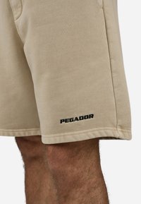 Pantalones cortos beige hechos de una tela suave, con un logo negro "PEGADOR" en la parte inferior izquierda y detalles de costura visibles en el dobladillo.