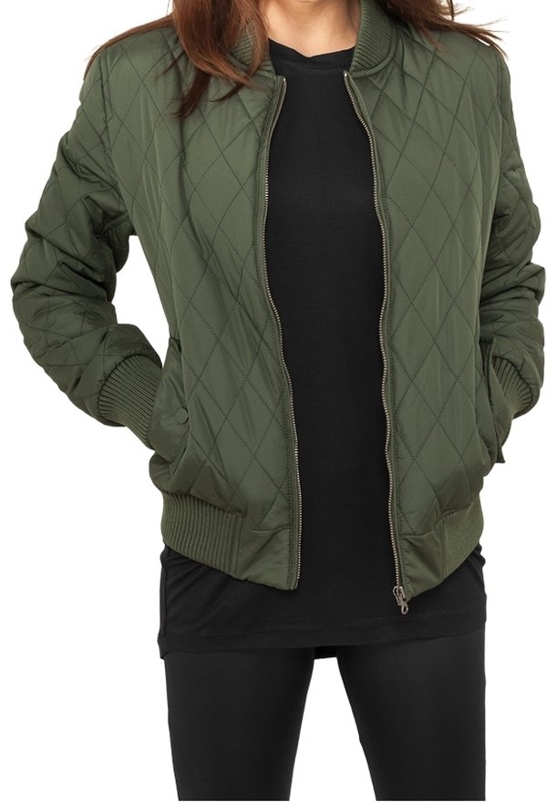 Femme portant une veste bomber matelassée vert olive dézippée sur un chemisier noir et un pantalon noir, les mains dans les poches de la veste.