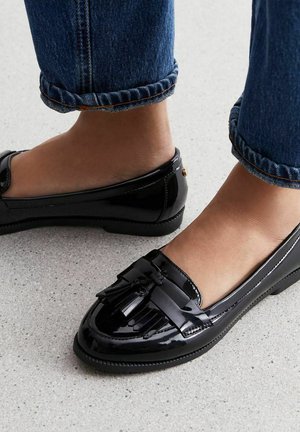 Black patent leather loafers with a flat sole, featuring decorative tassels on the toe and a smooth, shiny texture.