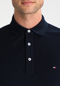 Homme portant un polo bleu marine avec trois boutons blancs et un petit logo rouge, blanc et bleu sur la poitrine.